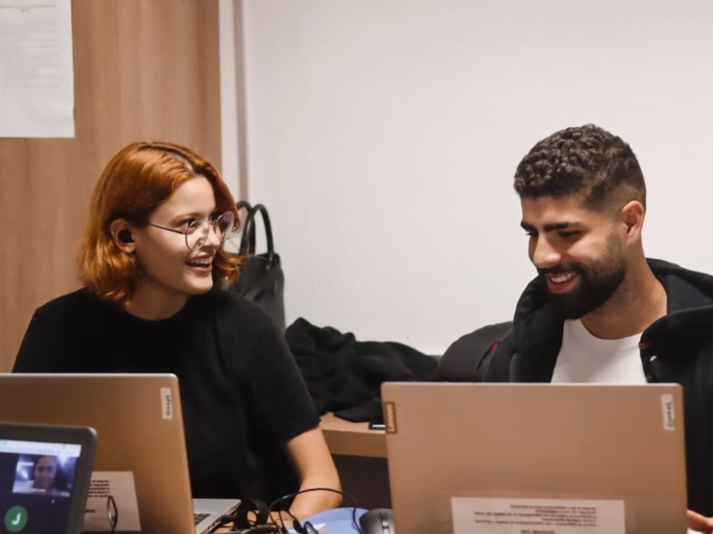 Dois estudantes conversam sentados em frente a dois computadores, no CESAR Summer Job.